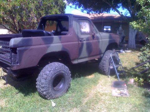 1990 Ford Bronco - This modified Ford Bronco, painted in camouflage, sits in a grassy yard with a spare tire nearby.