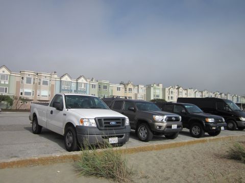 1991 Ford LTD Crown Victoria - A white Ford F-150 pickup truck is parked in a line with other vehicles, with colorful buildings in the background.