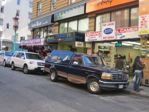 1991 Ford LTD Crown Victoria - A dark blue pickup truck with a camper shell is parked on a city street next to other vehicles and storefronts.