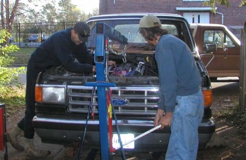 1988 Ford Bronco - Two men use a blue engine hoist to lower an engine into the front of a black Ford truck with the hood off, one leaning over the fender to g…