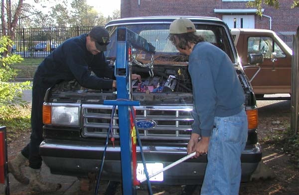 Two men lowering an engine into a black Ford truck with a blue engine hoist
