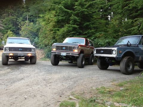 1984 Ford Bronco - Three SUVs, including a Ford Bronco, are parked on a dirt road surrounded by trees.