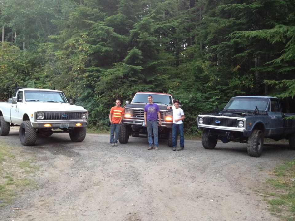 Three lifted pickup trucks parked on a dirt road with three people standing between them. The truck on the left is white, the middle truck is dark colored, and the truck on the right is dark blue with its hood open