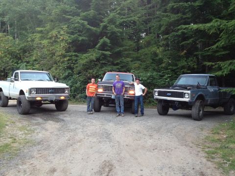 1984 Ford Bronco - Three lifted pickup trucks are parked on a dirt road with trees in the background.