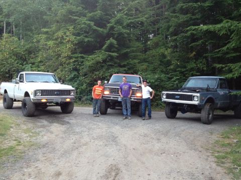 1984 Ford Bronco - Three pickup trucks are parked on a dirt road with a wooded background.