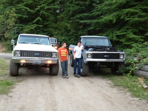 1984 Ford Bronco - Two lifted Chevrolet pickup trucks are parked on a dirt road with two men standing between them.