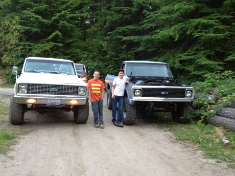 1984 Ford Bronco - Two lifted 1970s Chevrolet pickup trucks are parked on a dirt road with two people standing between them.