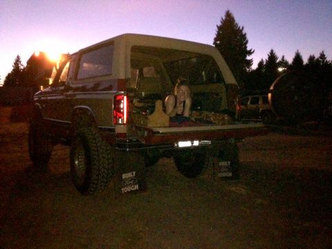 1984 Ford Bronco - A 1984 Ford Bronco is shown from the rear with its tailgate down, revealing a person relaxing in the cargo area as the sun sets.