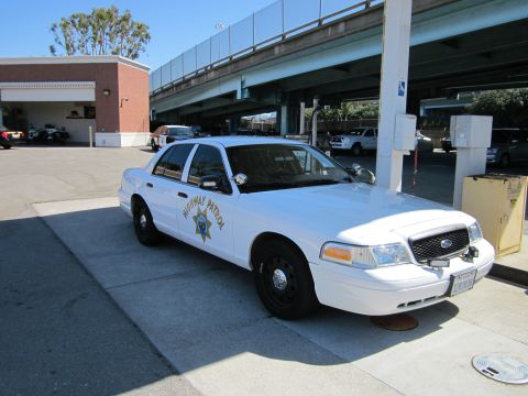 1991 Ford LTD Crown Victoria - A white Ford Crown Victoria police car is parked on a paved surface near a building and under an overpass.