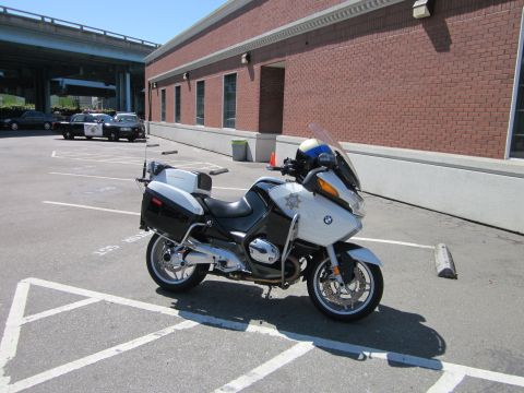1991 Ford LTD Crown Victoria - A white police motorcycle is parked in a lot, with two police cars visible in the background under an overpass.