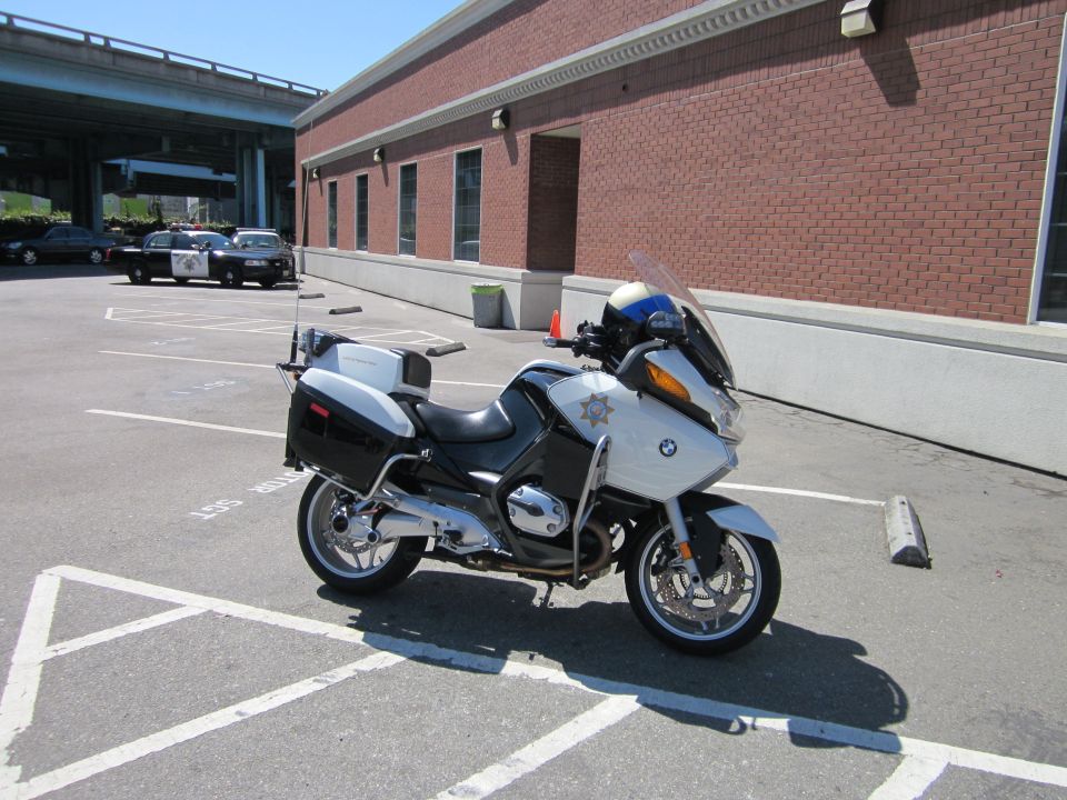 White police motorcycle parked in a lot, with police cars in the background