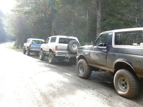 1991 Ford Bronco - Three lifted Ford 4x4s lined up along a gravel forest road, led in the foreground by a muddy dark blue Bronco with a white top, chrome-slot…