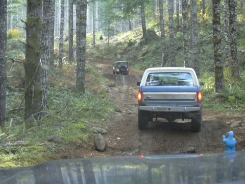 1991 Ford Bronco - A blue and white Ford Bronco navigates a muddy forest road, followed by another vehicle.
