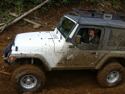 1991 Ford Bronco - A driver smiles from inside a white Jeep Wrangler heavily coated in mud while navigating an off-road trail.