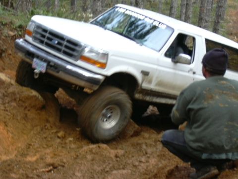 1991 Ford Bronco - A white 1990s Ford Bronco full-size SUV flexes through a deep red-clay rut on a wooded trail, front driver-side wheel drooped and mud-caked…