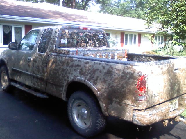 Mud-covered Ford F-150 extended-cab pickup, rear 3/4 view, loaded with firewood in driveway