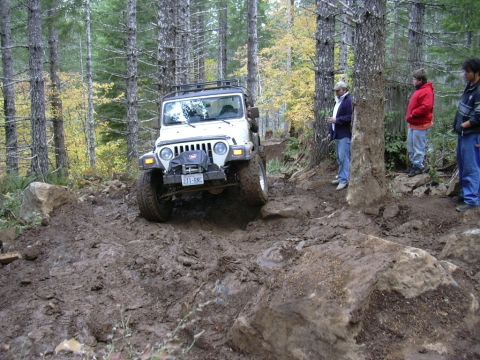 1991 Ford Bronco - A white Jeep Wrangler tackles a challenging muddy and rocky off-road trail in a wooded area.