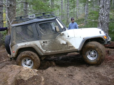 1991 Ford Bronco - A white Jeep Wrangler struggles through a muddy off-road course in a forest.