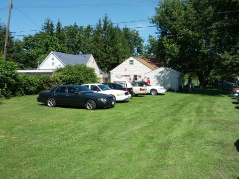 2007 Ford Crown Victoria - Several sedans are parked on a grassy area in front of residential buildings on a sunny day.