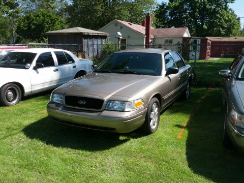 2007 Ford Crown Victoria - A tan 2007 Ford Crown Victoria is parked on a grassy lot, with other vehicles visible nearby.