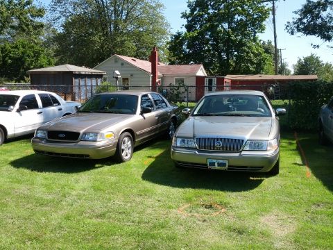 2007 Ford Crown Victoria - Two sedans are parked side-by-side on a grassy area in front of a residential building.