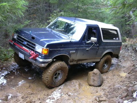 1991 Ford Bronco - A blue Ford Bronco navigates a challenging muddy off-road trail with snow patches.
