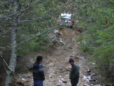 1991 Ford Bronco - A dark SUV navigates a challenging incline on a wooded trail, with two people observing from the side.
