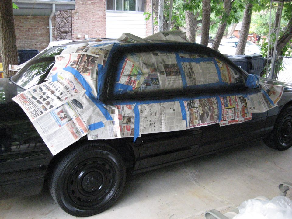Black Crown Victoria sedan masked with newspaper and blue tape for painting, parked under a carport