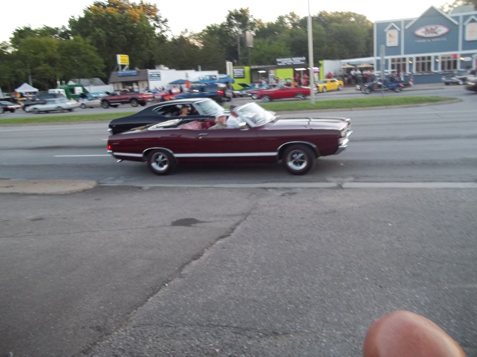 Maroon convertible with white interior and side trim driving on a street. Black car behind it