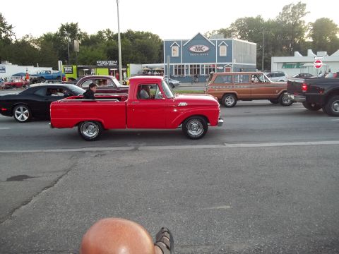 1970 Ford Ranchero - A red 1965 Ford F-100 pickup truck is seen driving down the road with other vehicles.