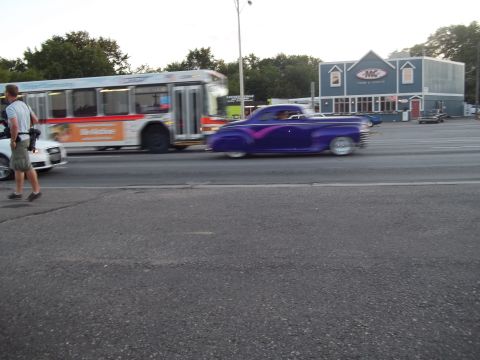 1970 Ford Ranchero - A purple customized coupe with pink racing stripes is seen driving on a street next to a bus.