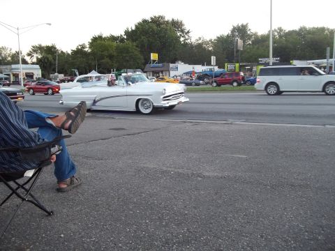 1970 Ford Ranchero - A white convertible cruises down the road, with other vehicles and spectators visible in the background.