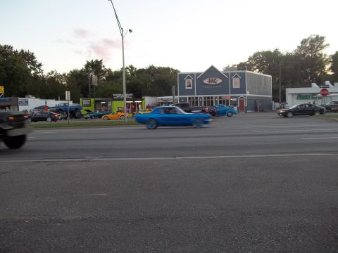 1970 Ford Ranchero - A blue 1967 Ford Mustang coupe is seen driving on a road, with other cars and buildings visible.