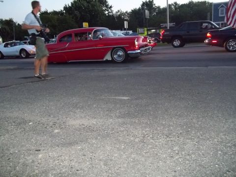 1970 Ford Ranchero - A man photographs a classic red sedan as it drives down the road, with other vehicles visible.
