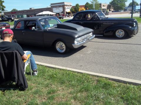 1970 Ford Ranchero - A man sits in a lawn chair watching two vintage cars drive by on a sunny day.