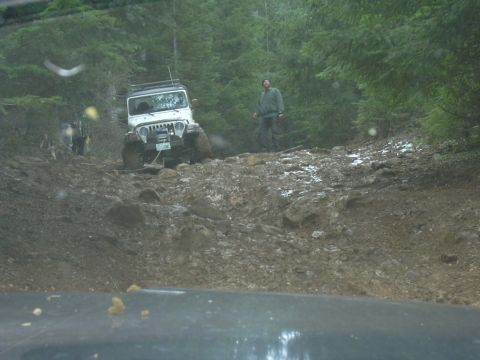 1991 Ford Bronco - A white Jeep Wrangler struggles on a steep, muddy, and rocky off-road path, with another vehicle partially visible behind it.