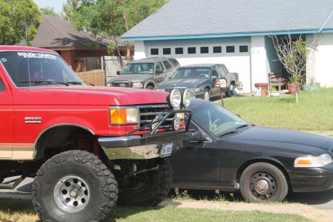 1990 Ford Bronco - A red Ford Bronco with large tires is parked next to a black sedan in front of a house.