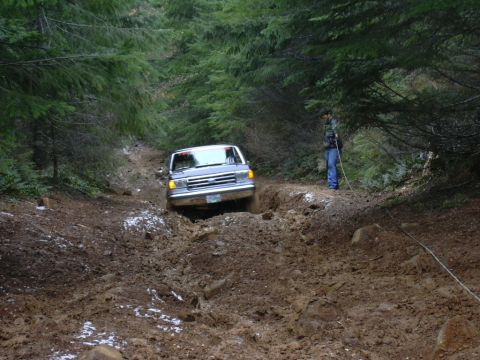 1991 Ford Bronco - A gray SUV drives through a muddy trail in a forest, with a person standing nearby.