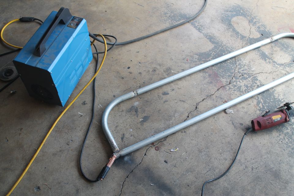 Blue welding machine with yellow and black cables next to bent metal tubing and a red grinder