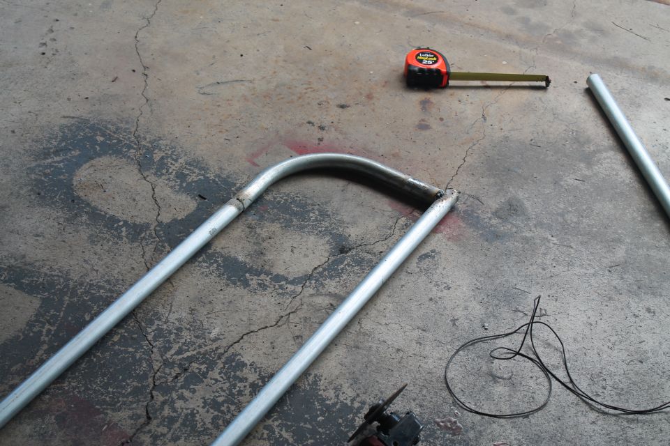 Metal tubing bent into a U-shape lies on a concrete floor next to a tape measure and wires
