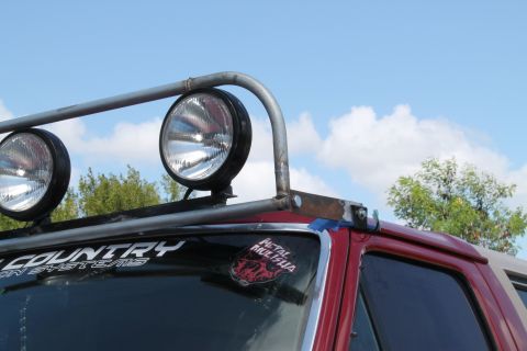 1990 Ford Bronco - Close-up of a red SUV's roof, showing a custom light bar with two large spotlights and various stickers on the windshield.