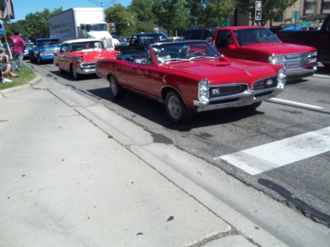 1970 Ford Ranchero - A line of cars, including a red convertible and a red pickup truck, are driving down a street on a sunny day.