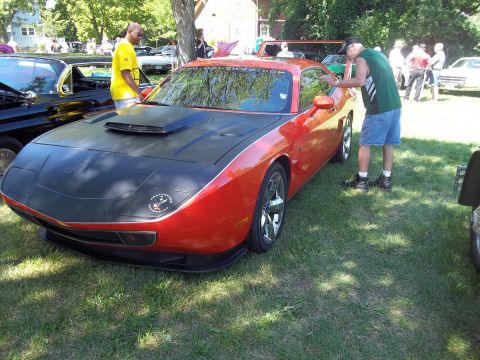 1970 Ford Ranchero - A man inspects an orange sports car with a black hood and scoop at an outdoor car show.
