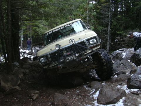 1991 Ford Bronco - A modified Ford Bronco navigates a challenging rocky terrain on a wooded trail, with a person watching nearby.