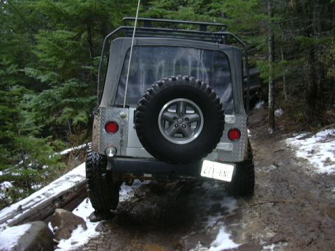 1991 Ford Bronco - A silver Jeep Wrangler navigates a muddy, snow-dusted trail through a forest.