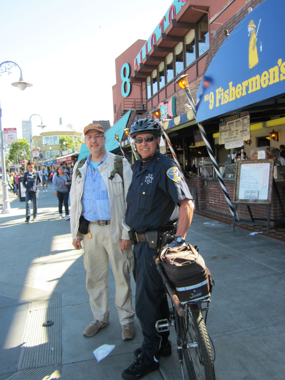 Two men, one in a police uniform, stand on a sidewalk next to a bicycle. A building with signage is behind them