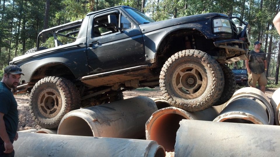 Modified black 1992 Ford Bronco with off-road tires driving over large concrete pipes. Another vehicle is visible in the background