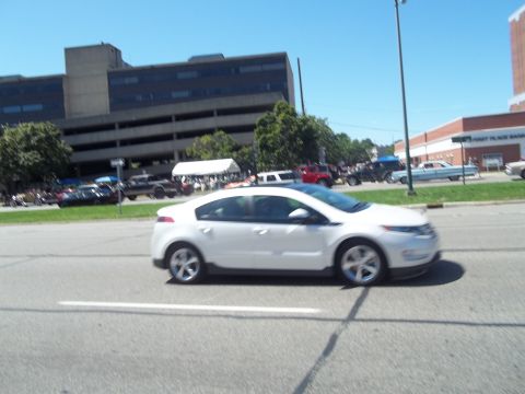 1970 Ford Ranchero - A white sedan is captured in motion on a sunny day, with a large building and other vehicles visible.