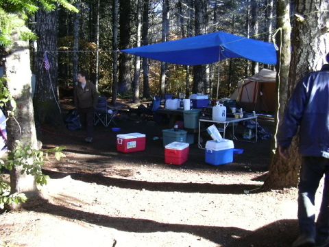 1991 Ford Bronco - A campsite is set up with a blue tarp providing shade over a table laden with supplies, surrounded by coolers and tents.