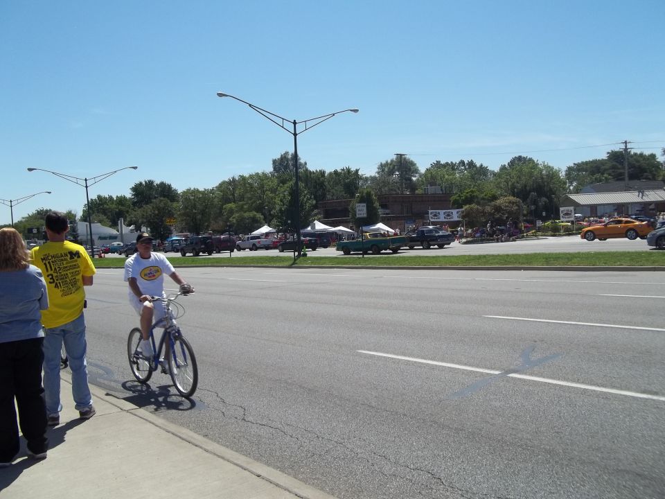 People on sidewalk watch cars at a show across the street. One guy rides a bike